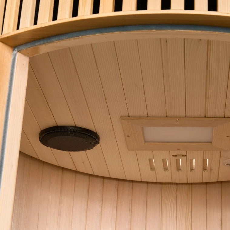 Close-up of the interior wooden ceiling of a Holl's Moon 1 wooden sauna cabin with a curved exterior with light natural wood planks, a round black vent or speaker, and a rectangular light fixture.