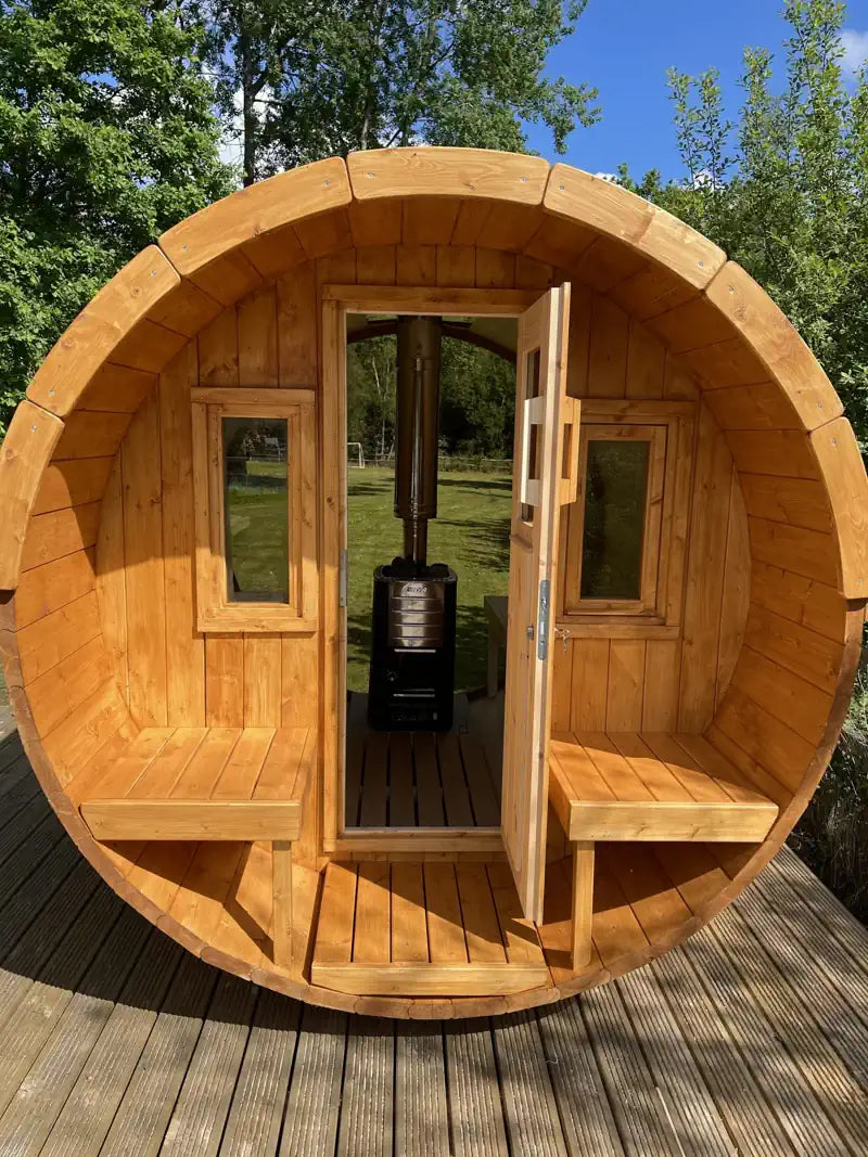 A round wooden 250 sauna cabin with built-in benches and an open door, photographed outdoors on a wooden deck with trees and blue sky in the background.