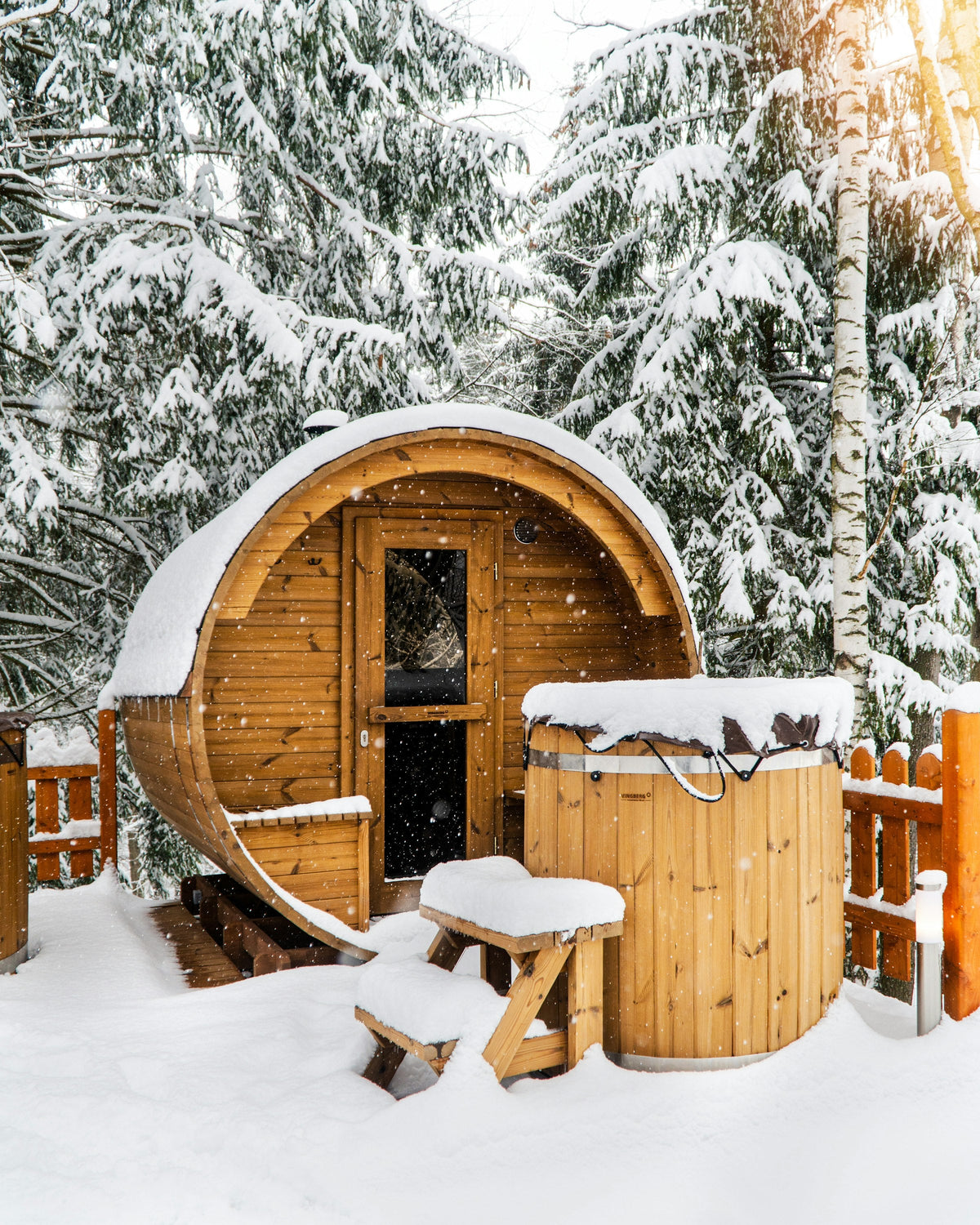 A barrel sauna and a timber tub under deep snow in a pine forest.