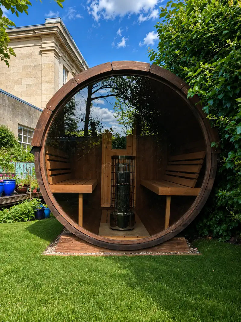 A round wooden 250 sauna cabin with built-in benches and a central heater, situated outdoors on a grassy lawn with a building and greenery in the background.