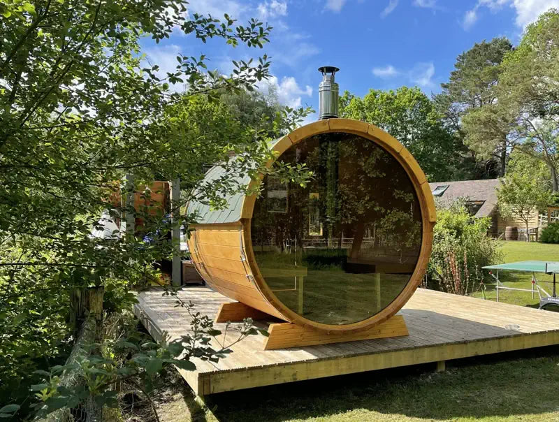 A round wooden 250 sauna cabin with large glass windows sits on a wooden platform in a backyard garden setting with trees and a blue sky.
