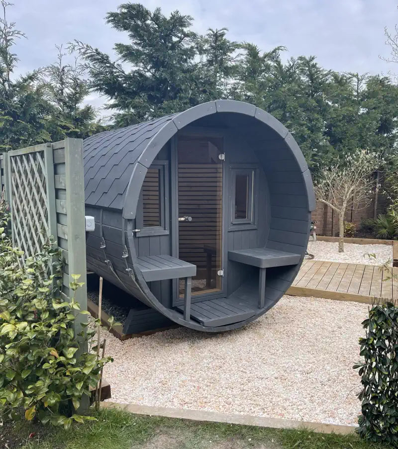 A dark gray wooden 250 sauna cabin with a circular opening, shown outdoors on a gravel surface with trees and shrubs in the background.