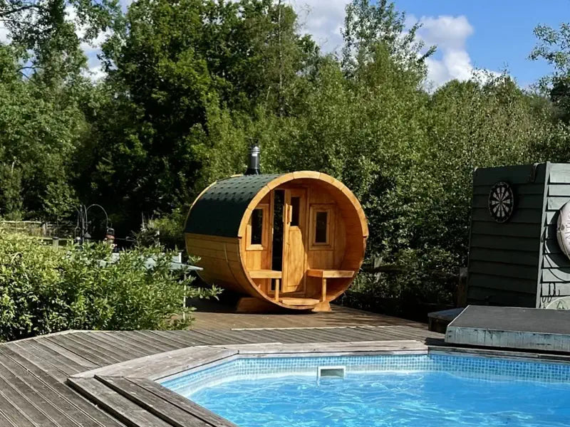 A wooden 250 sauna cabin with a dark green roof sits on a wooden deck beside a swimming pool, surrounded by trees and shrubs outdoors.