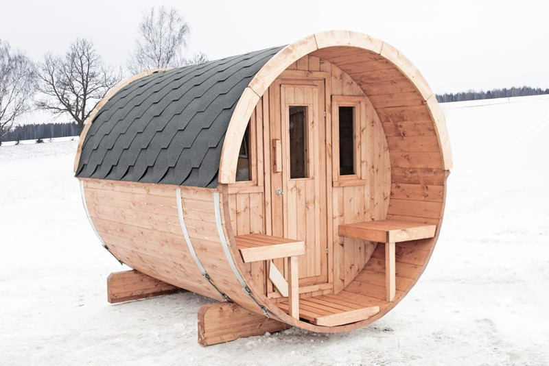 A wooden barrel-250 sauna cabin with a dark gray shingled roof, set outdoors in a snowy landscape.