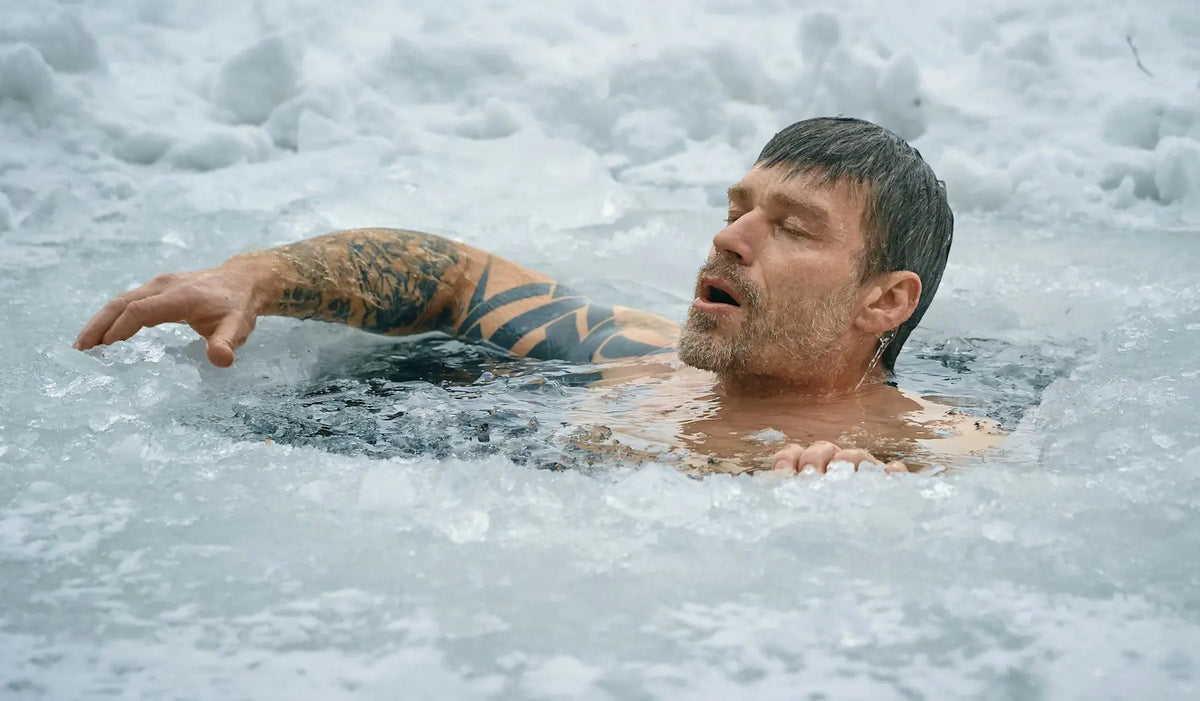 A man emerges from icy water, surrounded by snow and ice, with his face and arm visible above the surface.